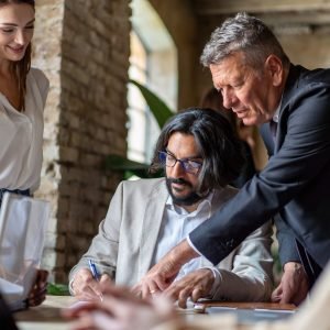 Diverse Team Signing a Professional Agreement in a Modern Office – business collaboration lifestyle concept Arab man signs a contract guided by an older man in a suit while a diverse group of people look on. Multicultural team works together to finalize a business deal, diversity and collaboration concept
